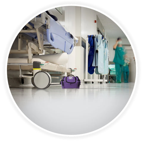 A contextual image showing the blue trauma bag placed on the floor of a clean hospital corridor, positioned next to a medical gurney with blurred healthcare professionals walking in the background.