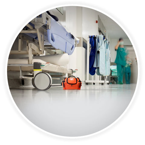 A contextual image showing the orange trauma bag placed on the floor of a clean hospital corridor, positioned next to a medical gurney with blurred healthcare professionals walking in the background.