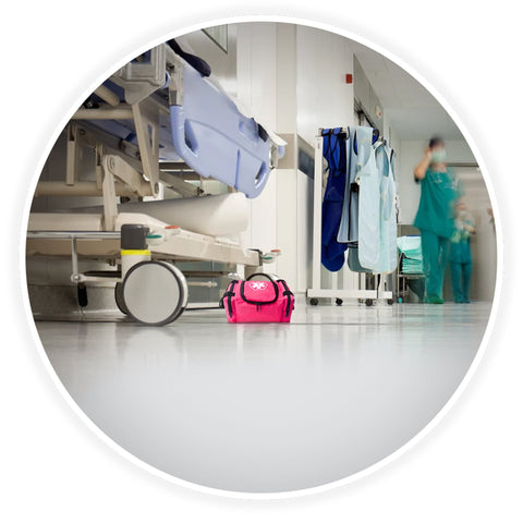 A contextual image showing the pink trauma bag placed on the floor of a clean hospital corridor, positioned next to a medical gurney with blurred healthcare professionals walking in the background.