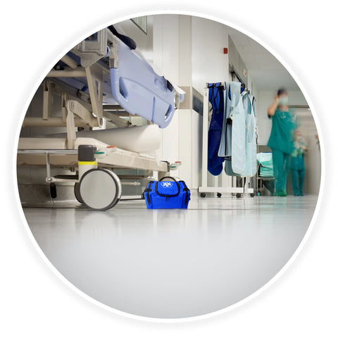A contextual image showing the blue trauma bag placed on the floor of a clean hospital corridor, positioned next to a medical gurney with blurred healthcare professionals walking in the background.