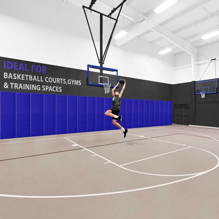 A basketball player jumping to dunk a ball in a gym where the wall behind the hoop is lined with a row of blue safety pads, accompanied by text reading "IDEAL FOR BASKETBALL COURTS, GYMS & TRAINING SPACES".
