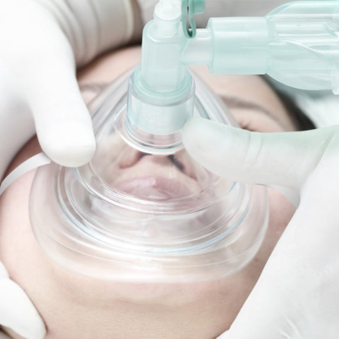 A close-up medical photograph showing a patient lying down with a clear anesthesia or oxygen mask covering their nose and mouth. Gloved hands are visible holding the mask securely in place to create a seal.