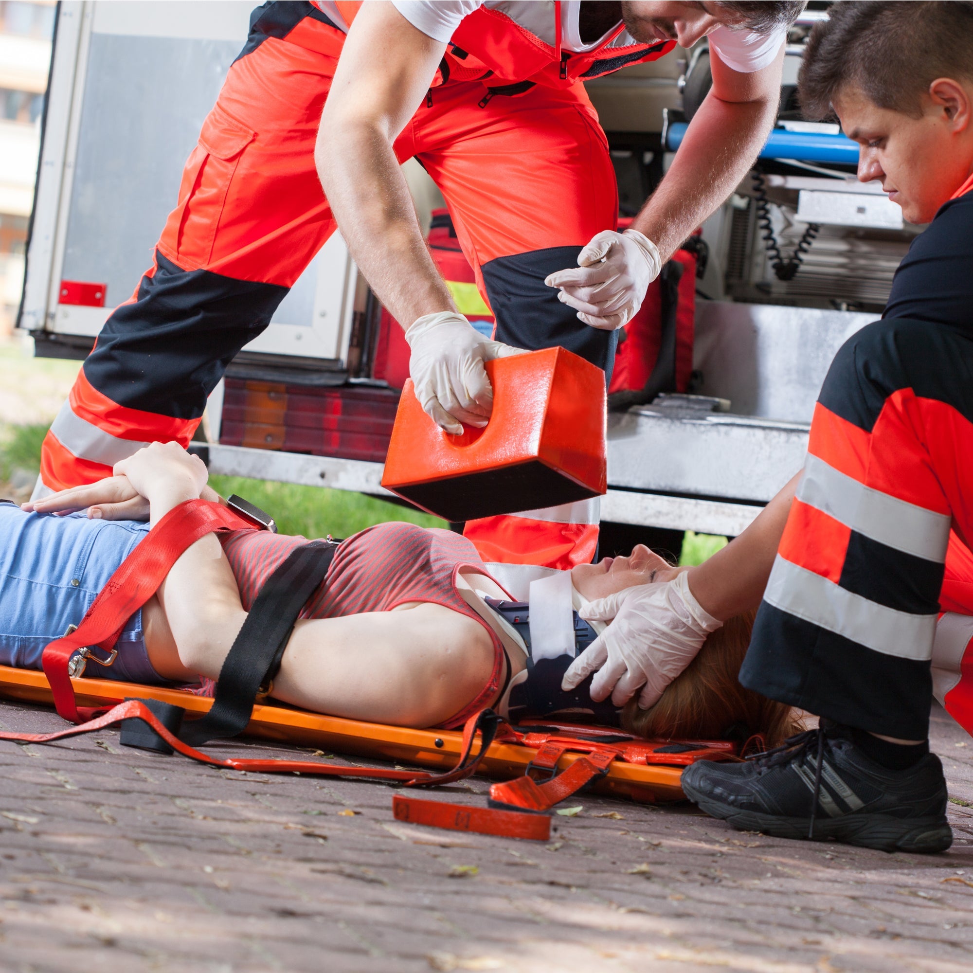 Two paramedics in high-visibility red and black uniforms attending to a patient strapped to an orange backboard. One responder is positioning an orange head immobilizer block while the patient wears a cervical collar.