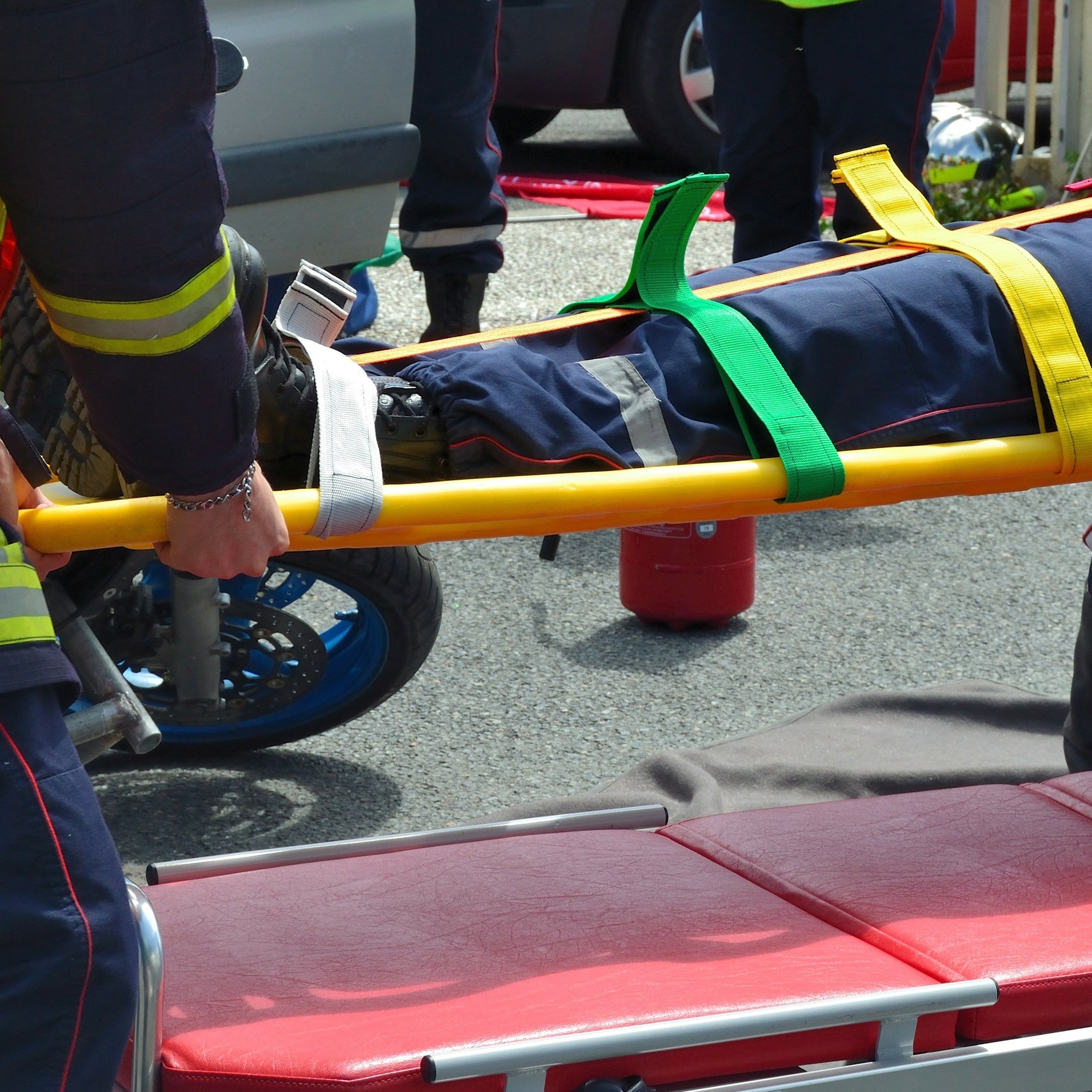 A close-up view of emergency responders lifting a patient secured to a yellow spine board. The image highlights the use of color-coded green and yellow patient restraint straps to keep the individual immobile during transport.