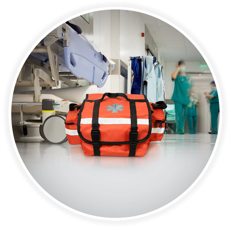 The orange trauma bag sits on a clean floor in a hospital hallway. The background is blurred but shows a medical gurney and doctors in scrubs walking by, establishing a professional medical setting.