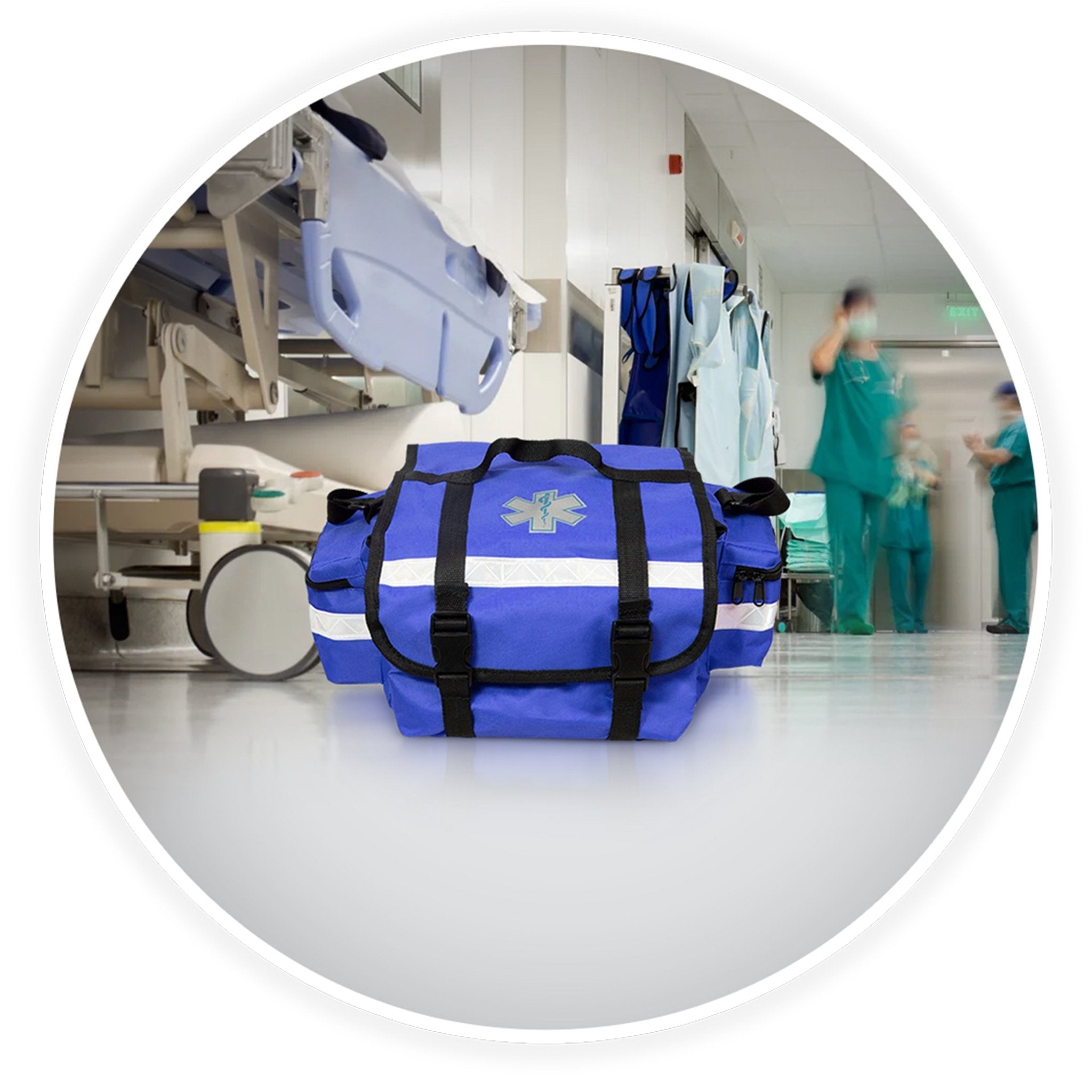 The blue trauma bag sits on a clean floor in a hospital hallway. The background is blurred but shows a medical gurney and doctors in scrubs walking by, establishing a professional medical setting.