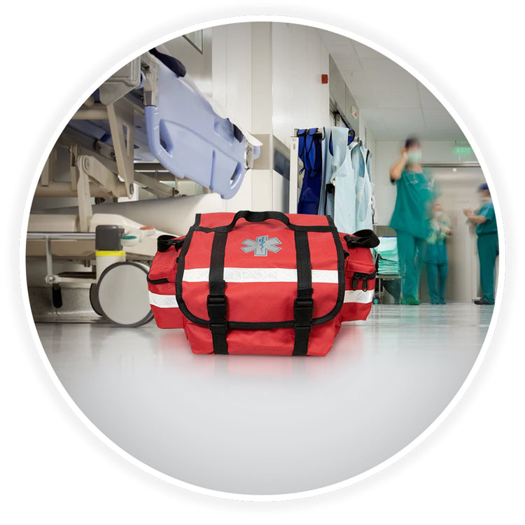 The red trauma bag sits on a clean floor in a hospital hallway. The background is blurred but shows a medical gurney and doctors in scrubs walking by, establishing a professional medical setting.