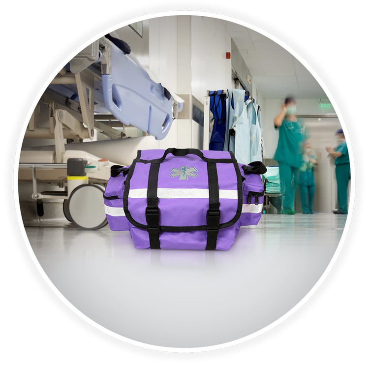 The purple trauma bag sits on a clean floor in a hospital hallway. The background is blurred but shows a medical gurney and doctors in scrubs walking by, establishing a professional medical setting.