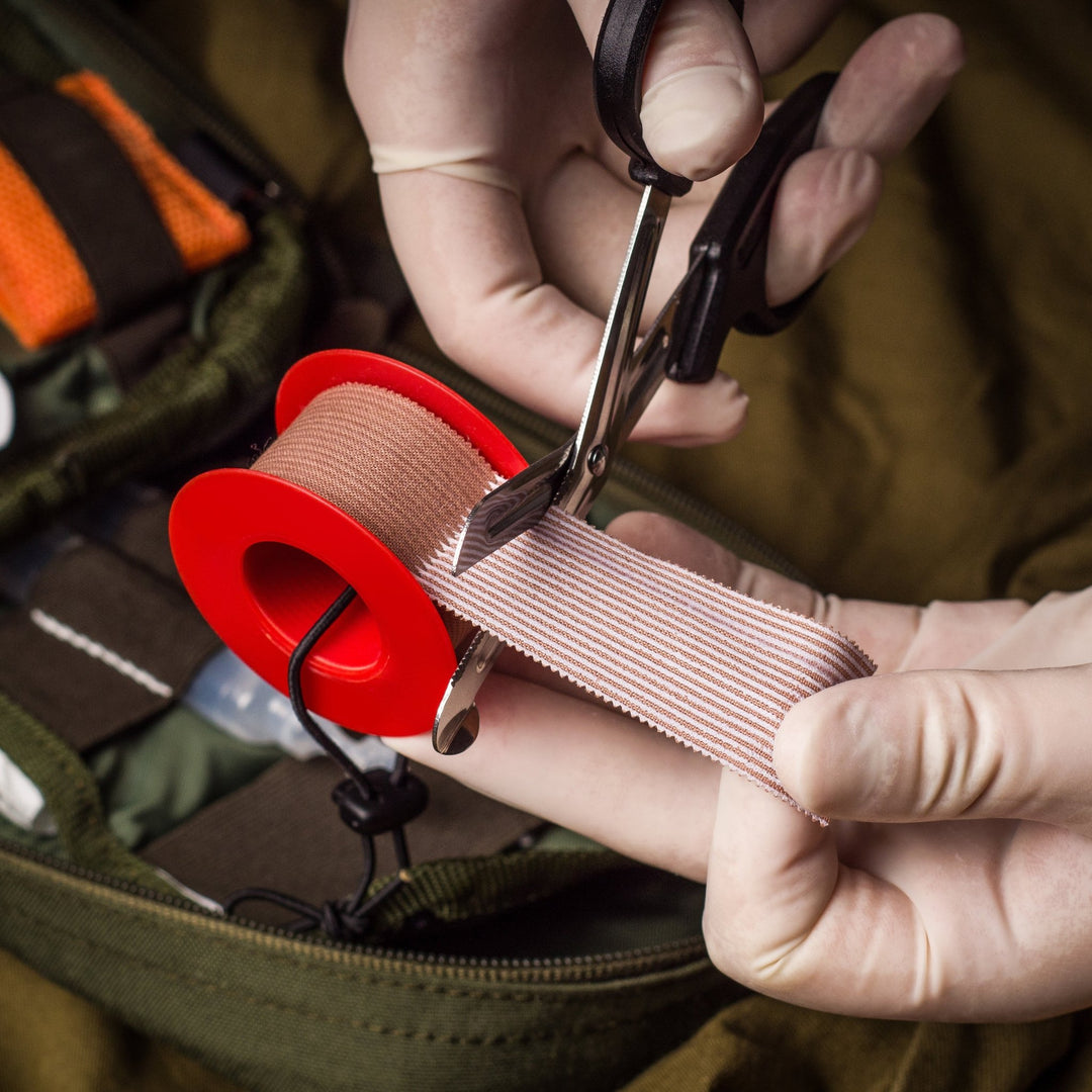 Hands wearing white latex gloves use the shears to cut a strip of elastic bandage. An open medical bag is visible in the background.