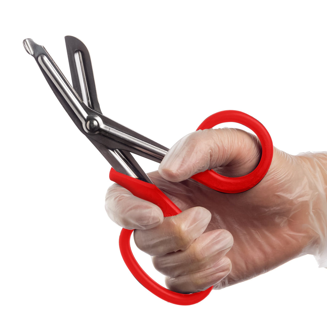 A hand wearing a clear medical glove holds a pair of open trauma shears with red handles against a white background.