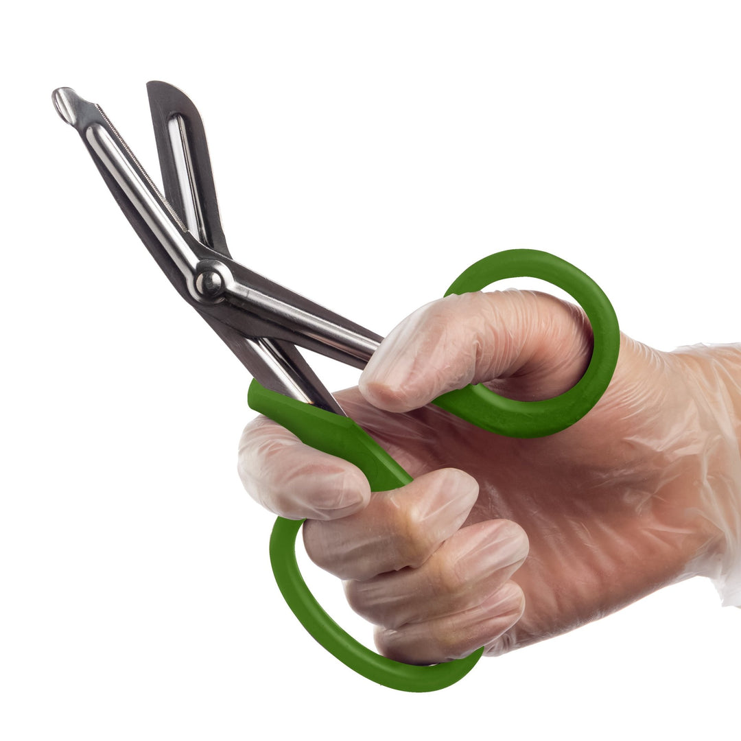 A hand wearing a clear medical glove holds a pair of open trauma shears with olive green handles against a white background.