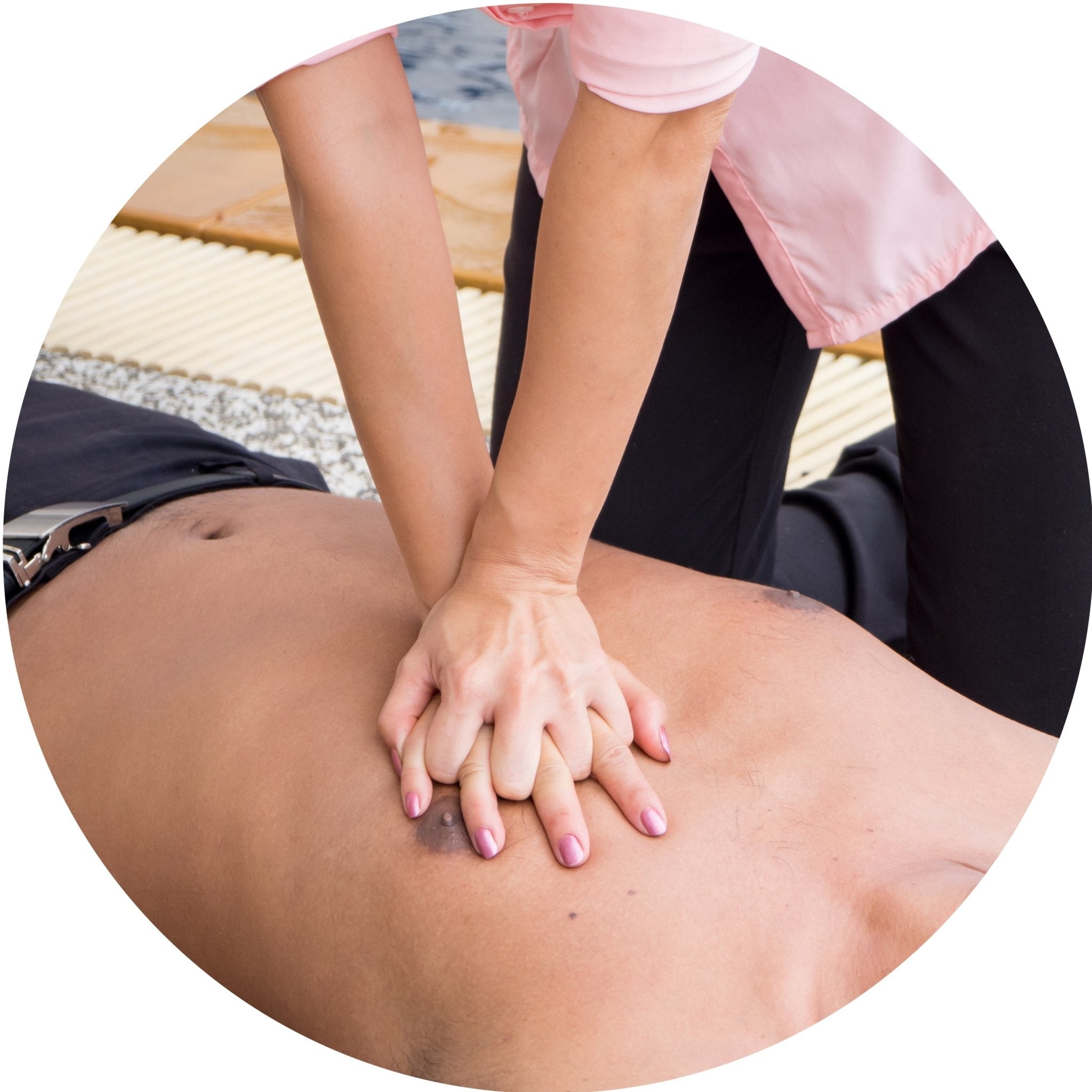A circular image showing a person in a pink shirt performing chest compressions on an unconscious, bare-chested man, demonstrating the correct hand placement for CPR. The hands are interlaced and positioned in the center of the man's chest.