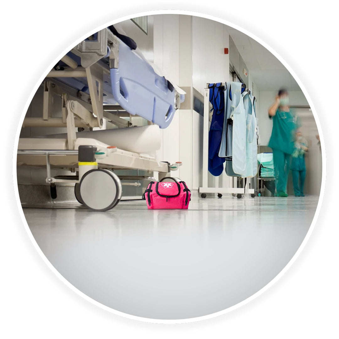 A contextual image showing the pink trauma bag placed on the floor of a clean hospital corridor, positioned next to a medical gurney with blurred healthcare professionals walking in the background.