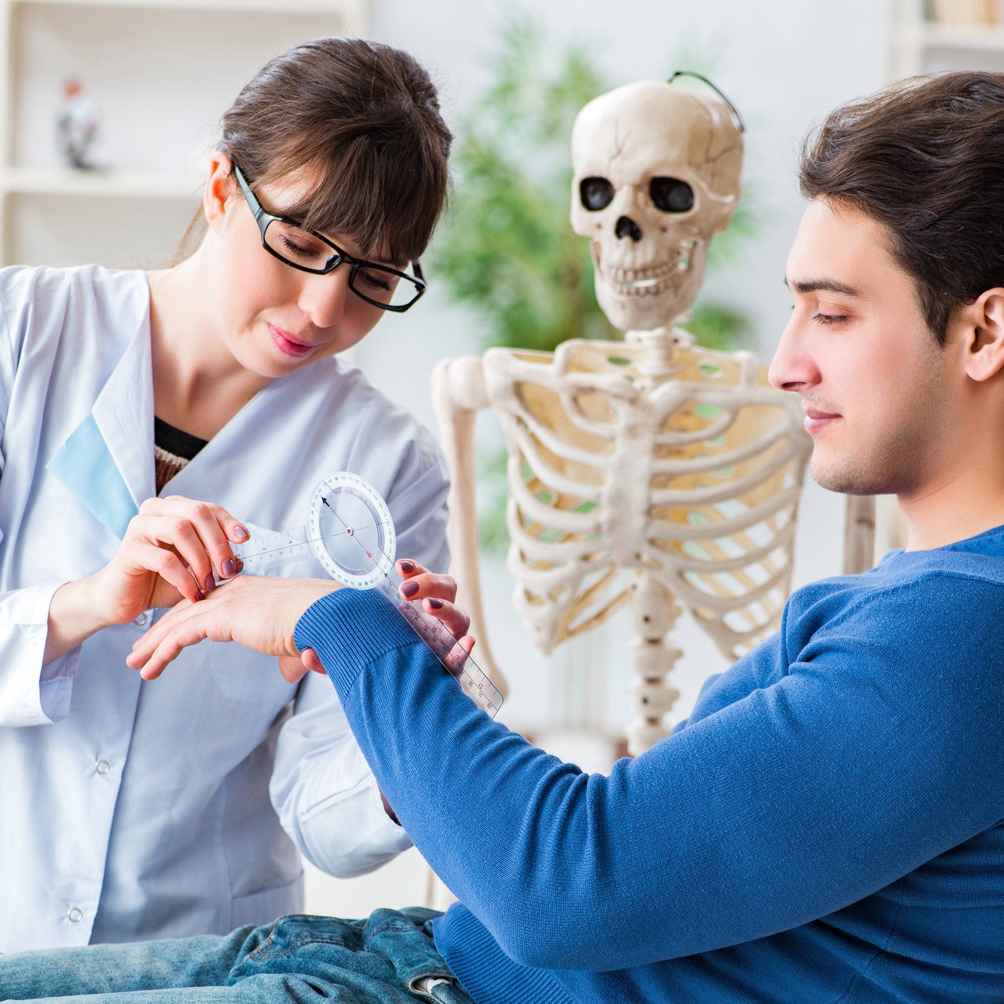 A female medical professional using a clear goniometer to measure the range of motion of a male patient's wrist, with a human skeleton model visible in the background.