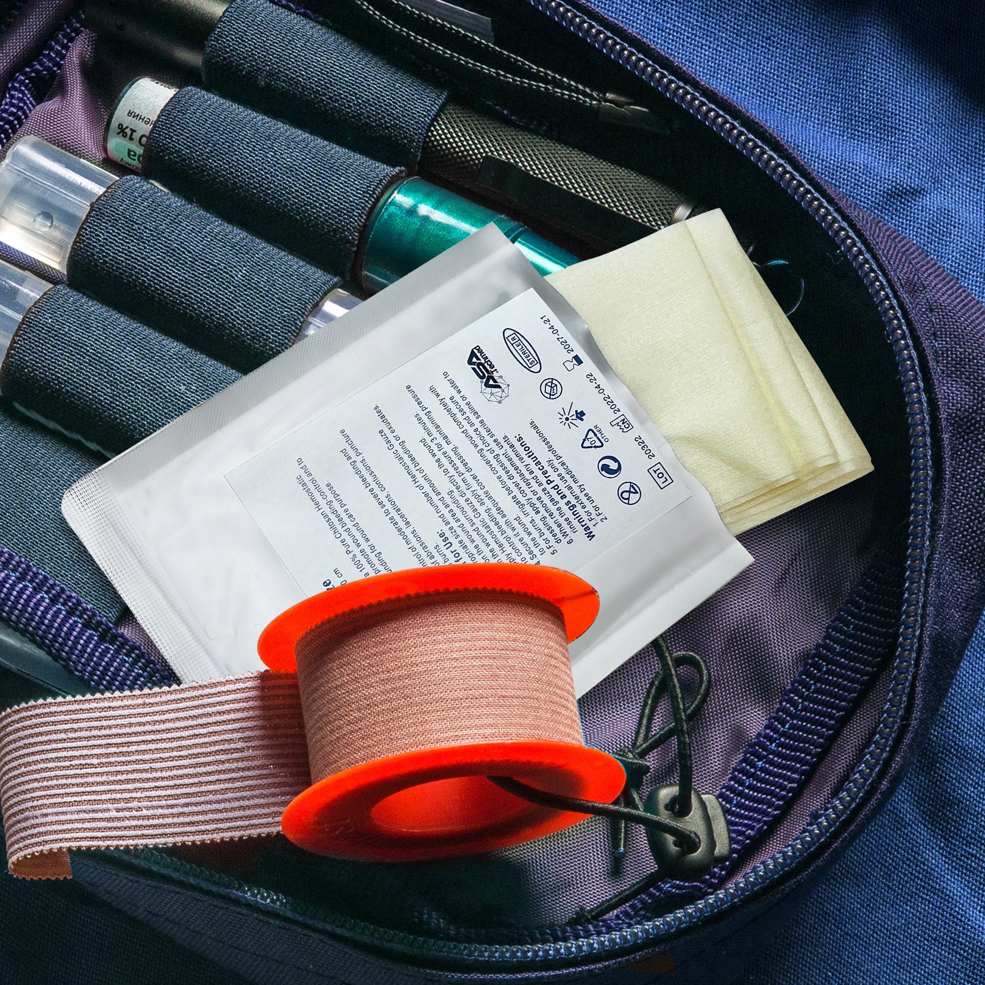 A hemostatic gauze packet stored inside an open tactical first aid bag, resting on top of an orange medical tape roll and adjacent to other emergency supplies.