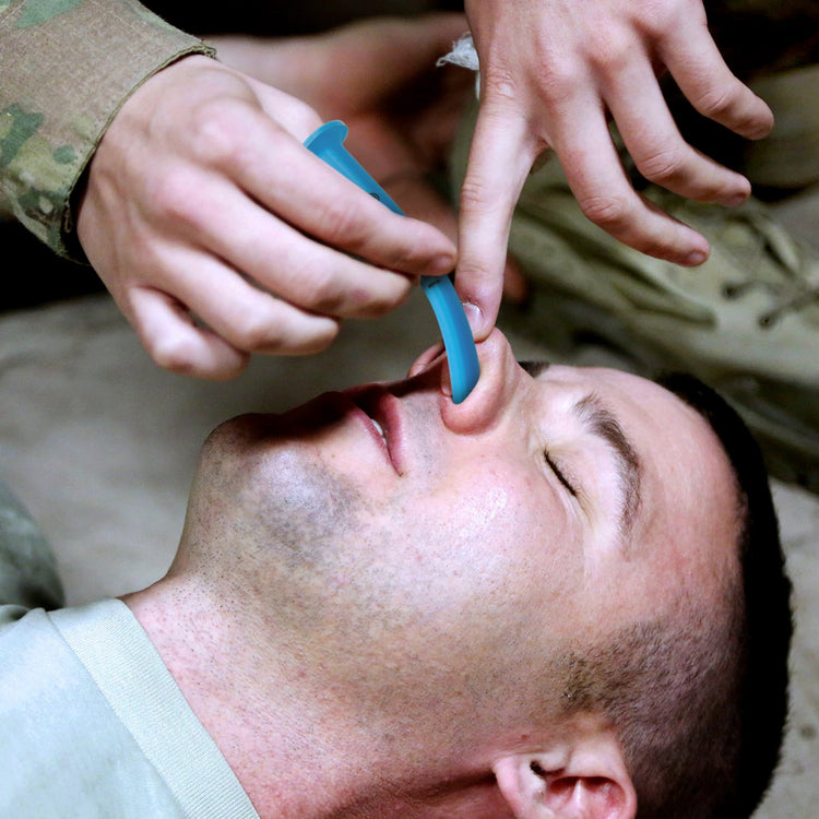 A realistic field application scene. A person in military camouflage fatigues is shown carefully inserting the blue Nasopharyngeal Airway into a patient's nostril to secure their airway.