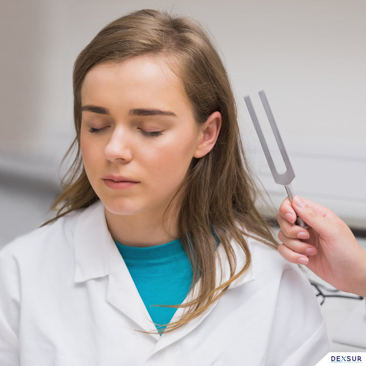 A medical professional in a clinical setting holds a silver C512 tuning fork near the ear of a seated female patient whose eyes are closed. The professional is conducting a diagnostic hearing or vibration test.