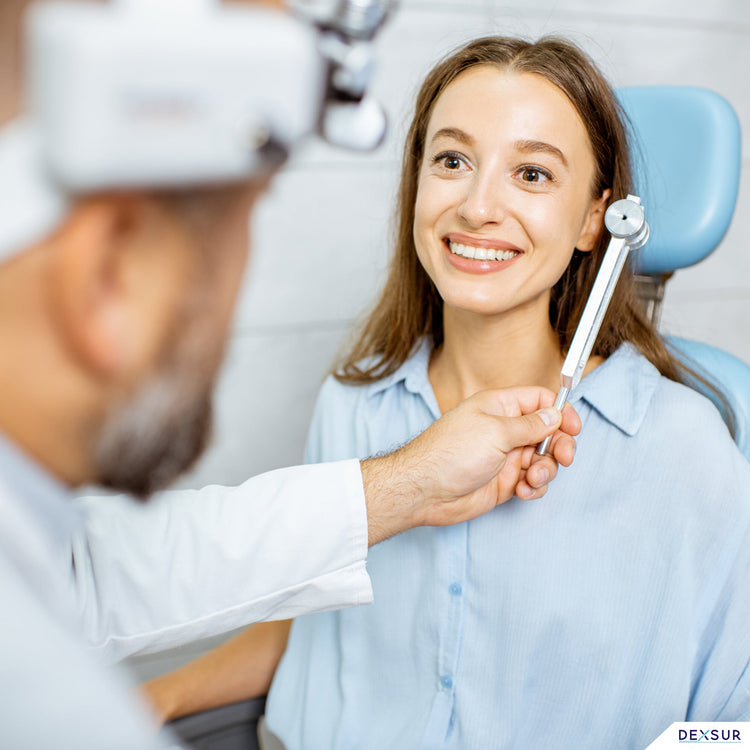 A lifestyle image of a medical professional performing a hearing or bone conduction test on a smiling female patient. The professional is holding a silver C256 tuning fork near the patient's ear in a clinical office setting.