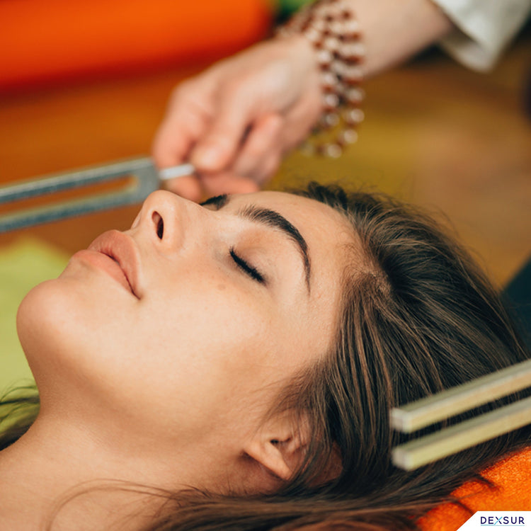 A close-up lifestyle image of a woman lying down with her eyes closed, receiving a sound therapy or Reiki treatment. A practitioner holds two vibrating silver tuning forks near the sides of the woman's head.