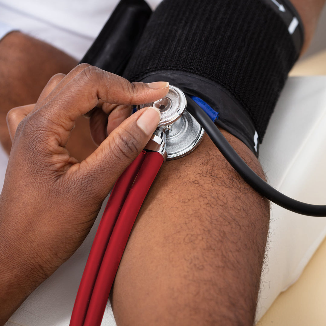 A close-up lifestyle image of a medical professional using the red dual-head stethoscope to monitor a patient's blood pressure. The stethoscope chest piece is held firmly against the patient's arm just below a black blood pressure cuff.