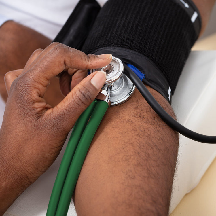 A close-up lifestyle image of a medical professional using the green dual-head stethoscope to monitor a patient's blood pressure. The stethoscope chest piece is held firmly against the patient's arm just below a black blood pressure cuff.