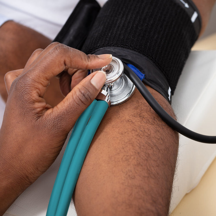 A close-up lifestyle image of a medical professional using the teal dual-head stethoscope to monitor a patient's blood pressure. The stethoscope chest piece is held firmly against the patient's arm just below a black blood pressure cuff.