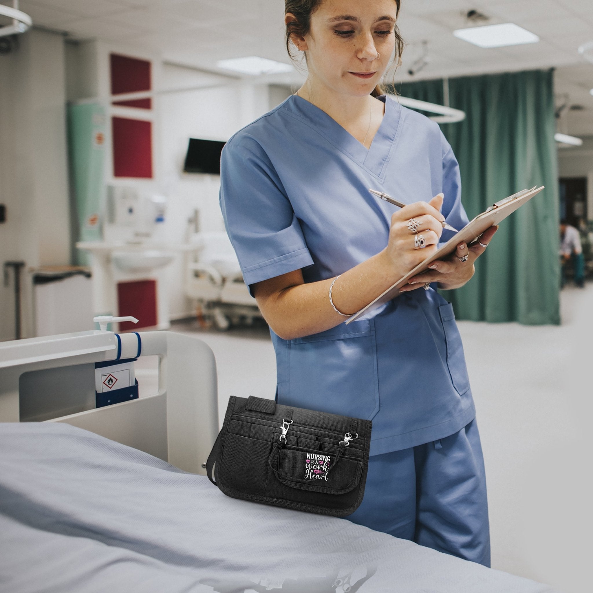 A medical professional in blue scrubs wearing the black nursing utility belt while taking notes on a clipboard in a hospital setting.