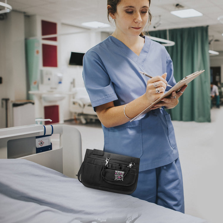 A medical professional in blue scrubs wearing the black nursing utility belt while taking notes on a clipboard in a hospital setting.