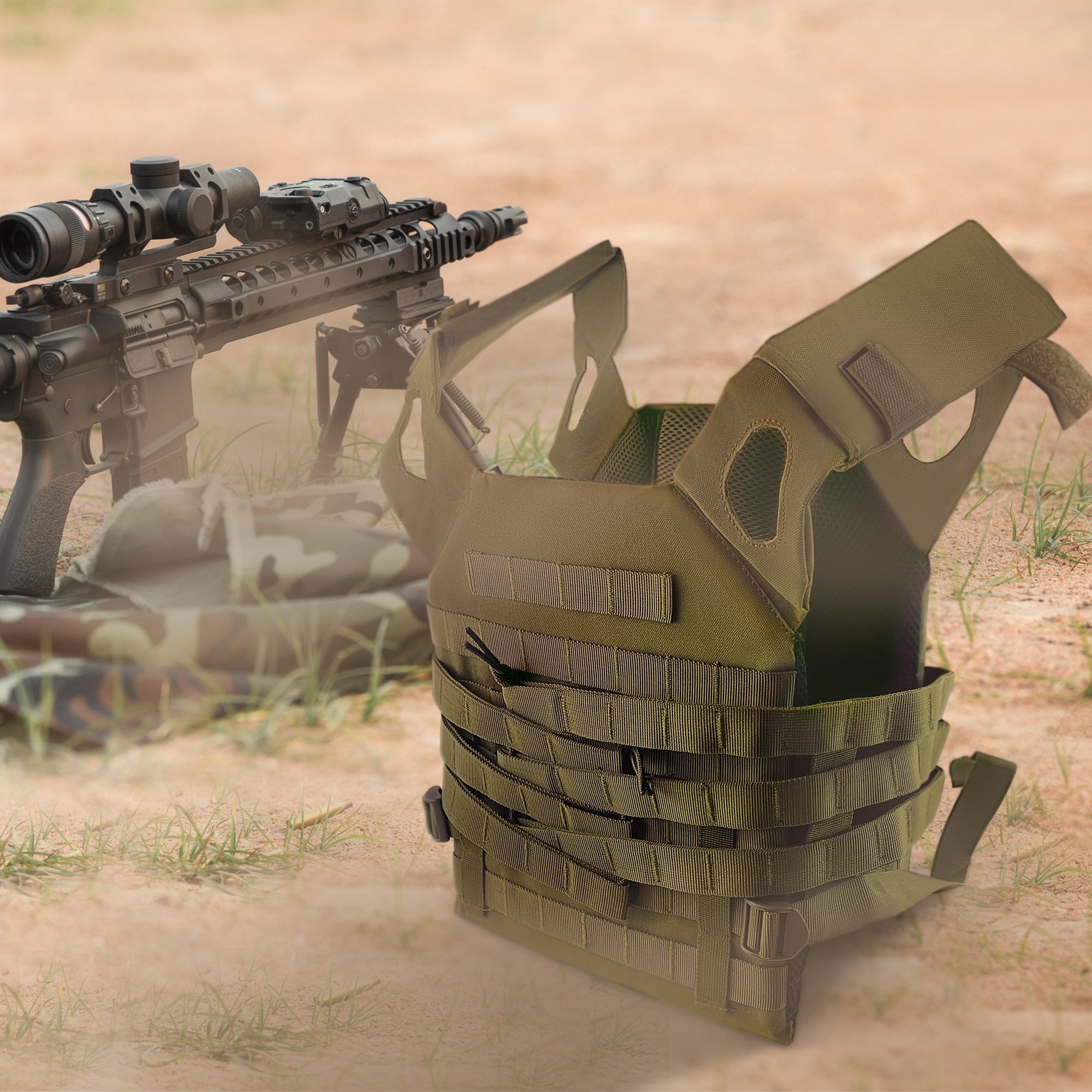 An olive green tactical plate carrier vest sitting on the ground in a sandy outdoor environment. In the background, a black tactical rifle equipped with a scope and bipod rests next to the vest.
