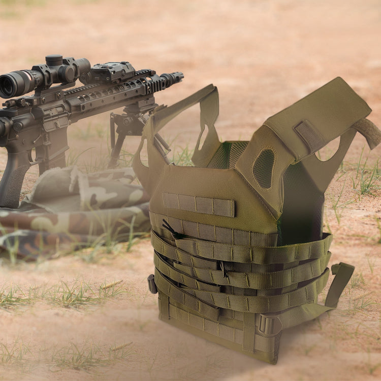 An olive green tactical plate carrier vest sitting on the ground in a sandy outdoor environment. In the background, a black tactical rifle equipped with a scope and bipod rests next to the vest.