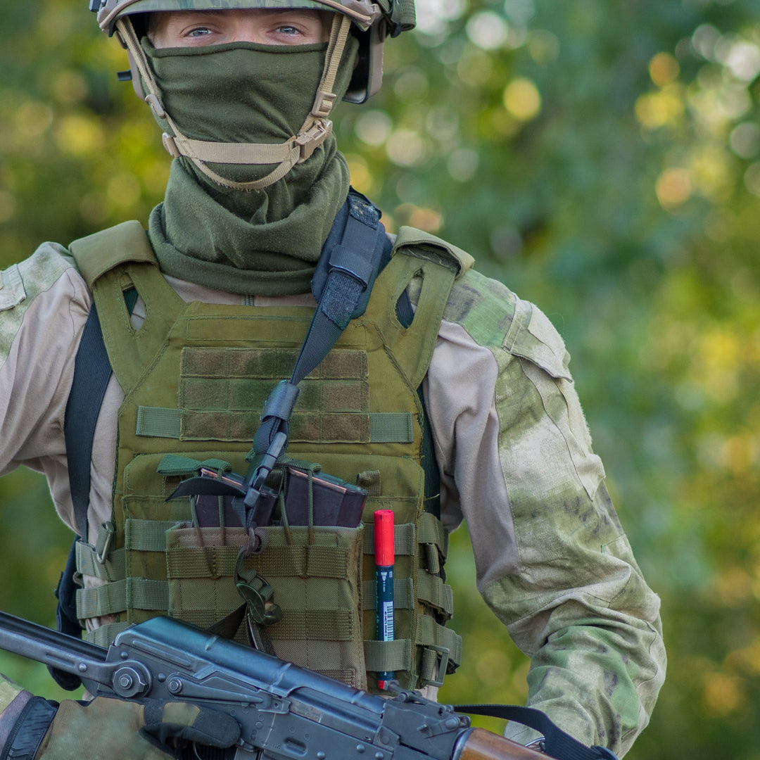 A soldier wearing full tactical gear, including an olive green plate carrier vest loaded with magazines and a red marker. The individual is wearing a matching green balaclava and helmet while holding a rifle in a wooded outdoor setting.
