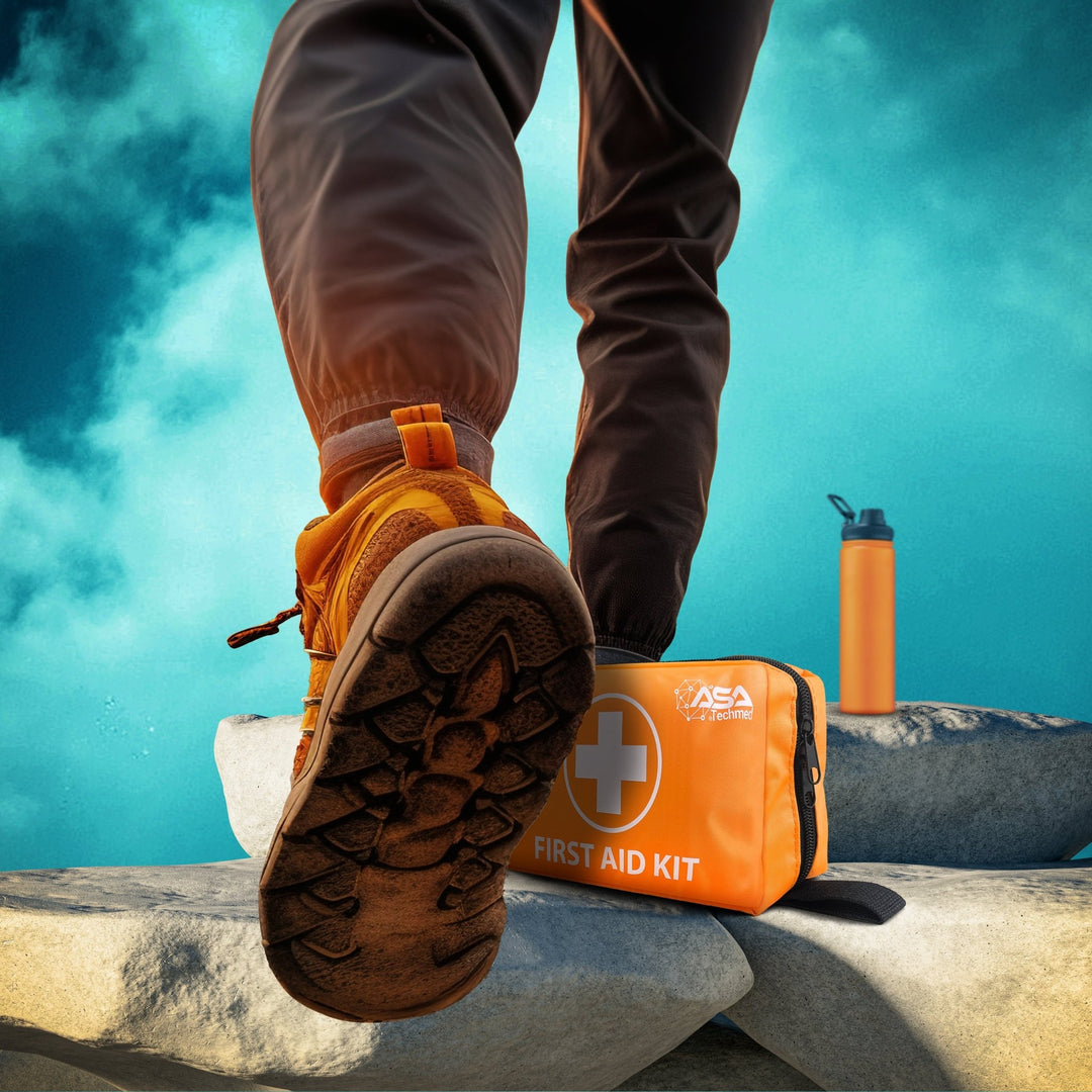 A low-angle lifestyle shot focusing on the hiking boots of a person stepping onto large rocks against a blue sky. Next to their feet rests the orange "ASA Techmed" first aid kit and an orange water bottle.